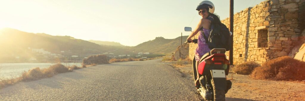 Woman riding scooter on scenic Greek road at sunrise, capturing adventure and travel vibes.