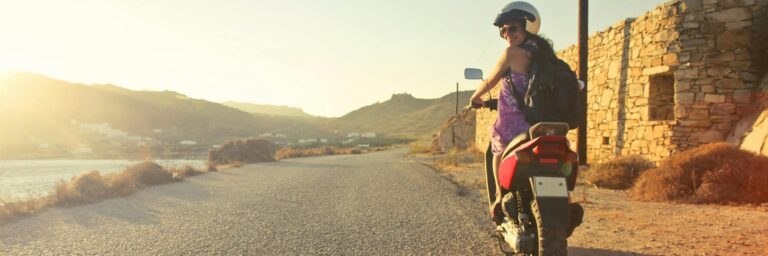 Woman riding scooter on scenic Greek road at sunrise, capturing adventure and travel vibes.
