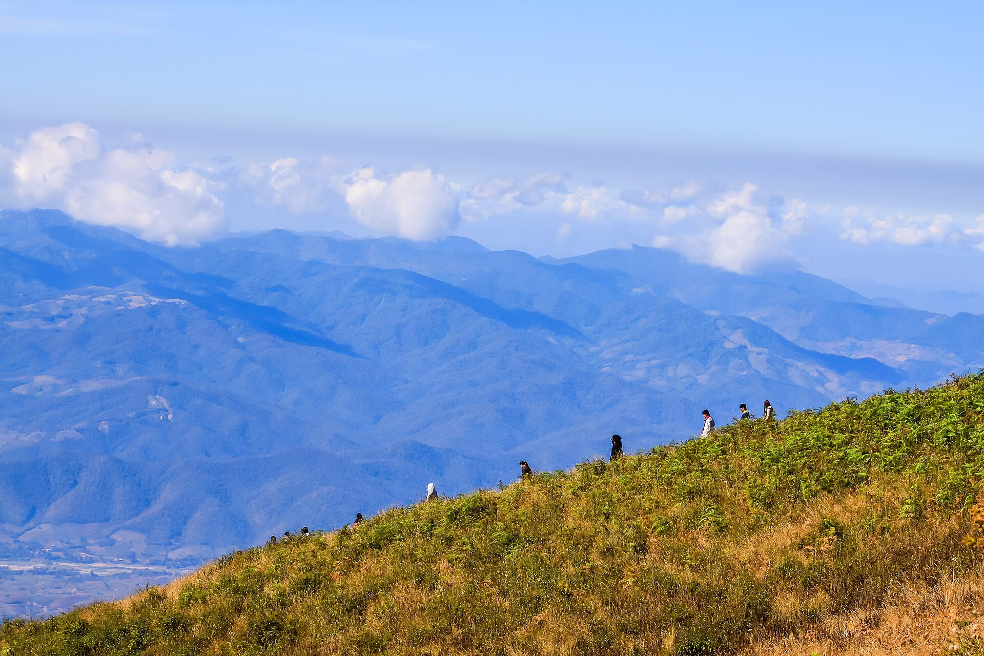 Walkers on the grassy Kew Mae Pan ridge on Doi Inthanon under a clear sky, with blue mountain ranges below